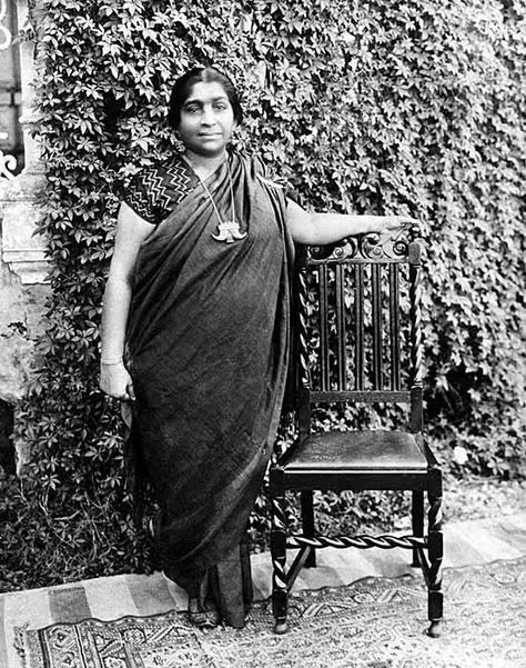 Full-length photograph of Sarojini Naidu standing beside a carved wooden chair in front of an ivy-covered wall.