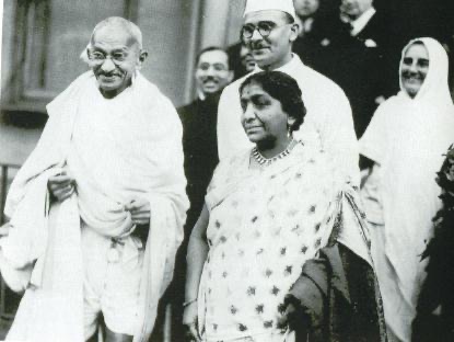 Gandhi in a white shawl standing beside Sarojini Naidu, with other members of a delegation behind them, in London.